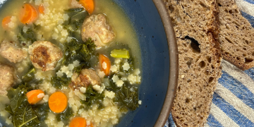Italian Wedding soup with carrots, kale, meatballs, and tiny pasta in a blue bowl with a blue striped towel and bread on the side